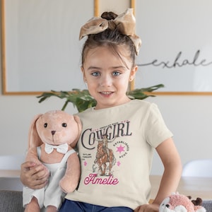 May include: A young girl wearing a cream colored t-shirt with a vintage-style graphic of a cowgirl riding a horse. The shirt says "Cowgirl" in a black script font and "American Made" and "Established 1907" in smaller font. The girl's name, "Amelie", is printed in pink below the graphic.