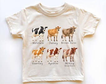 May include: A cream-colored short-sleeve t-shirt featuring a graphic print of six different cow breeds. The cows are labeled Holstein, Jersey, Brown Swiss, Guernsey, Ayrshire, and Milking Shorthorn in black text below each respective illustration.