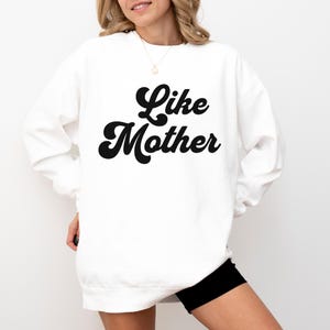May include: White sweatshirt with the text "Like Mother" in a bold, black, retro font. The sweatshirt has long sleeves and a crew neck. The overall style is casual and comfortable.