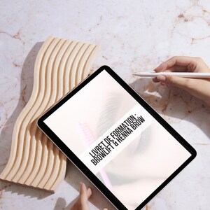 Puede incluir: Una tableta blanca con un borde negro que muestra un documento titulado "LIVRET DE FORMATION: BROWLIFT & HENNA BROW". La tableta está sobre una superficie de madera marrón claro.