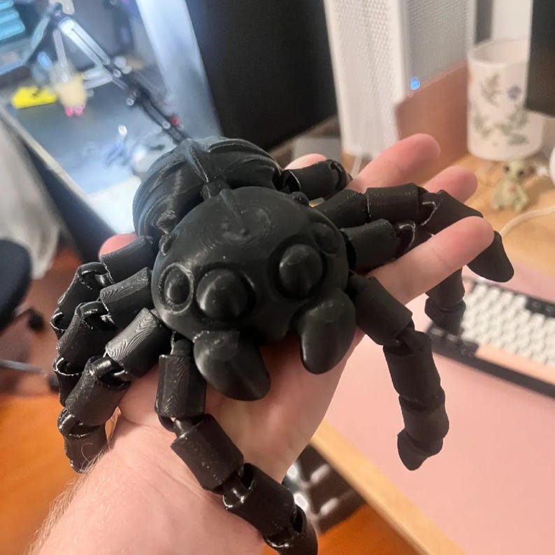May include: A black, articulated spider toy with a segmented body and legs. The spider has a rounded head and body, with multiple eyes visible. The legs are jointed, allowing for movement. The toy is held in a hand.