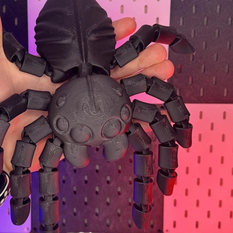 May include: A black articulated spider toy with segmented legs and body. The toy is made of plastic and has a detailed, textured surface. The spider's legs are poseable, allowing for various positions. The toy is held in a hand.