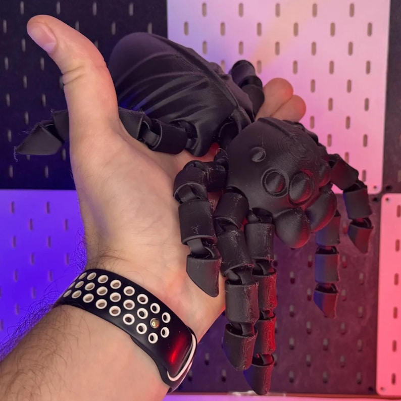 May include: A black articulated spider toy with multiple legs and a segmented body. The toy is held in a hand, showcasing its size and detail. The spider's design features a realistic appearance, with a focus on its intricate structure and dark color.