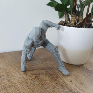 K&ouml;nnte beinhalten: Graue Figur eines Superhelden in dynamischer Pose, mit detailliertem Panzerdesign. Die Figur kniet mit einem ausgestreckten Arm, was auf Action hindeutet. Die Figur steht auf einer Holzoberfl&auml;che.