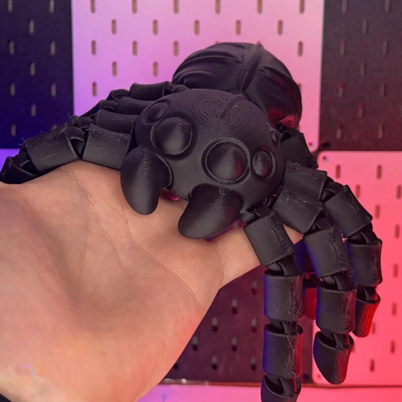 May include: A black articulated spider toy with a segmented body and legs. The spider has a detailed head with multiple eyes and a textured surface. The legs are poseable, allowing for various positions. The toy is held in a hand.