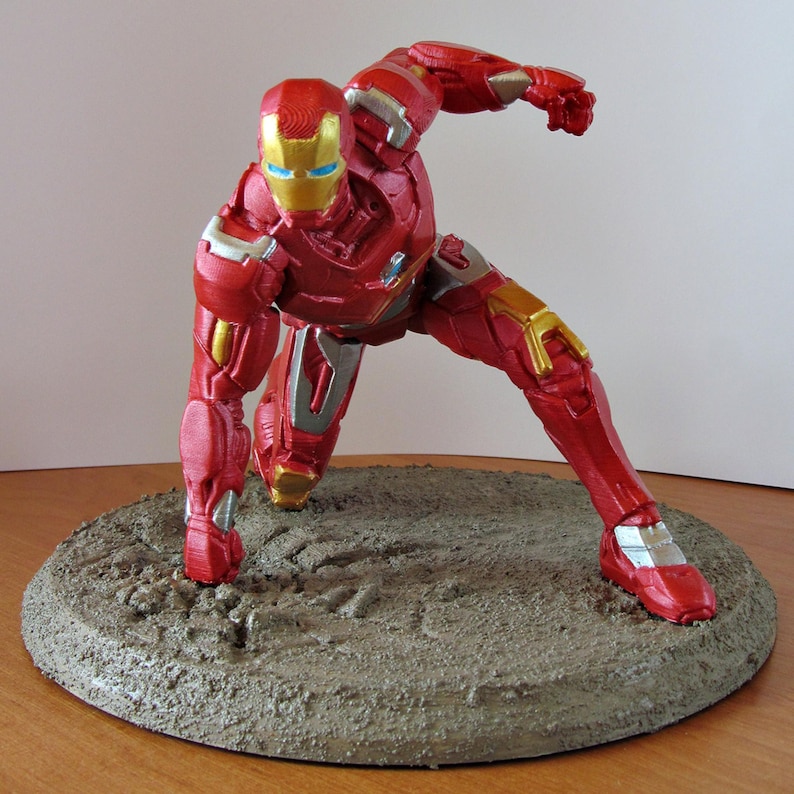 K&ouml;nnte beinhalten: Eine rot-goldene Iron Man-Figur in einer dynamischen Pose, platziert auf einer strukturierten, kreisf&ouml;rmigen Basis. Die Figur hat einen goldenen Helm mit blauen Augen und silbernen und goldenen Akzenten. Die Figur ist etwa 20 cm gro&szlig;.