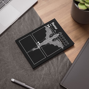 May include: A black notepad with a white dragon illustration and the word "Review" at the top. The notepad has sections for "Thoughts" and "Tropes", with a star rating system. The notepad is on a wooden desk.