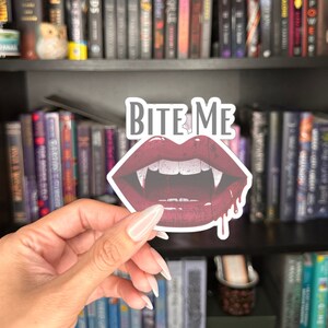 May include: A sticker featuring a pair of red lips with fangs and the words "Bite Me" in gray. The lips have a dripping blood effect. The sticker is held in front of a bookshelf filled with books.