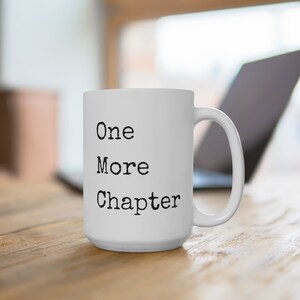 Könnte beinhalten: Weißer Keramikbecher mit dem schwarzen Text "One More Chapter", ideal für Leser. Der Becher hat einen großen Henkel und steht auf einer Holzoberfläche, mit einem Laptop im Hintergrund.
