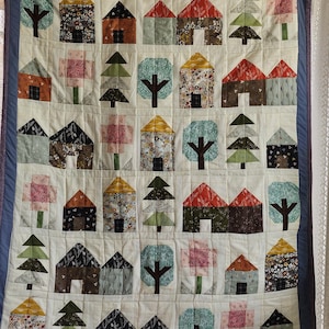 May include: A white quilt with a blue border features a patchwork design of houses, trees, and flowers. The quilt is made up of various fabrics in shades of brown, green, red, and yellow.