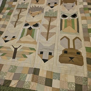 May include: A handmade quilt with a patchwork design featuring animal faces, such as a fox, rabbit, and owl. The quilt has a cream background with green, brown, and tan accents. The border is a green fabric.