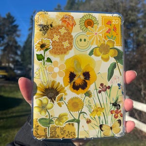 May include: A clear phone case with a yellow floral design. The design includes sunflowers, bees, butterflies, and a smiley face. The background has a yellow and white checkered pattern. The case is held up against a sunny, outdoor background.