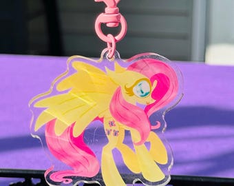 Puede incluir: Un llavero de acrílico transparente con un personaje de poni amarillo con una larga melena y cola rosa, grandes ojos azules y alas. El personaje tiene un símbolo de mariposa en el flanco. Está unido a un cierre giratorio de metal rosa en forma de corazón.