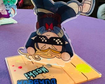 Puede incluir: Una figura de acrílico transparente con un personaje de dibujos animados en posición de manos. La figura está sobre una base rectangular con las palabras "Dungeon Mesh" en azul y verde. La base tiene salpicaduras de pintura amarilla y roja.
