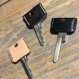 Puede incluir: Tres llaves con fundas de cuero. Dos fundas son negras y una es de color beige. Las fundas negras tienen costuras blancas y la funda beige tiene costuras negras. Las llaves están sobre una superficie de madera.
