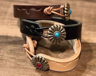 Puede incluir: Tres pulseras de cuero con detalles metálicos decorativos. La pulsera superior es de cuero marrón con una piedra turquesa, la pulsera del medio es de cuero negro con una piedra turquesa y la pulsera inferior es de cuero marrón claro con una piedra roja.