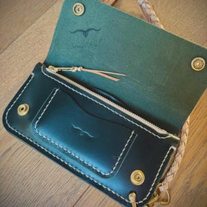 Puede incluir: Cartera de cuero verde oscuro abierta, con cierre de cremallera y ranuras para tarjetas. La cartera tiene costuras blancas, herrajes de latón y una correa de cuero trenzado. El interior es de un verde más claro y el texto "Savage Soul" es visible.
