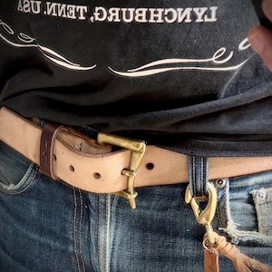 May include: A natural tan leather belt with a brass buckle and a dark brown leather keeper loop. The belt is worn over blue denim jeans. A brass swivel hook with a braided leather tassel is attached to a belt loop on the jeans.