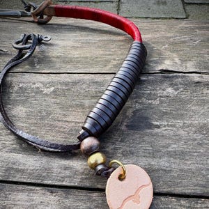 May include: Handmade leather necklace with a red wrapped handle and a dark gray, layered design. The necklace features a brown leather strap, a light brown pendant with a bird design, and brass and wooden beads.