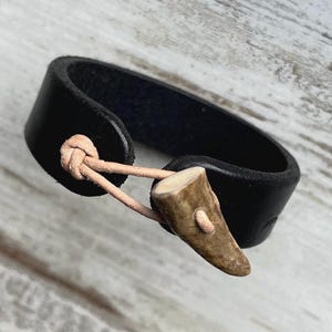 May include: A black leather bracelet with a natural antler clasp. The bracelet is a simple band design, secured with a light brown leather cord and a small, light brown antler piece. The bracelet is likely for everyday wear.