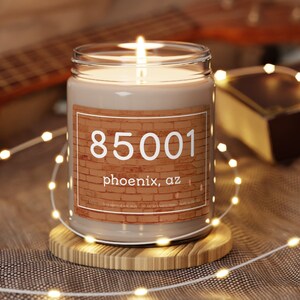May include: A lit candle in a clear glass jar with a label featuring the number "85001" and "phoenix, az" on a brick background. The candle sits on a wooden coaster, with string lights in the foreground.