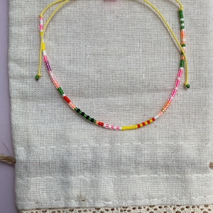 May include: A colorful beaded bracelet with a yellow string and pink knot. The bracelet is on a white fabric background.