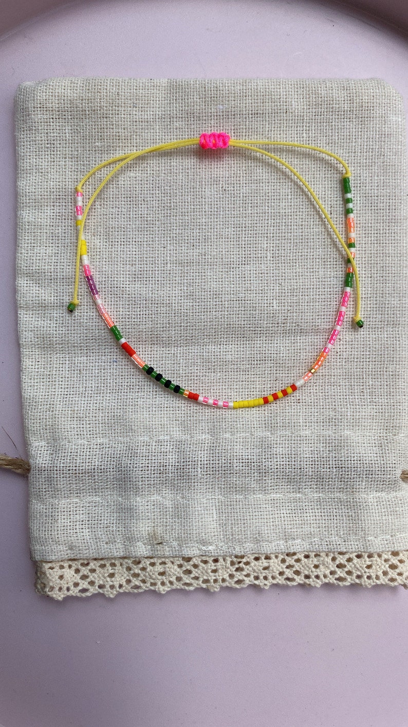 May include: A colorful beaded bracelet with a yellow string and pink knot. The bracelet is on a white fabric background.
