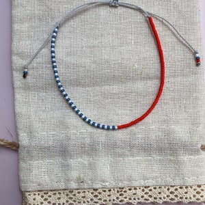 May include: A red and blue beaded bracelet with a white string. The bracelet is on a white fabric background.
