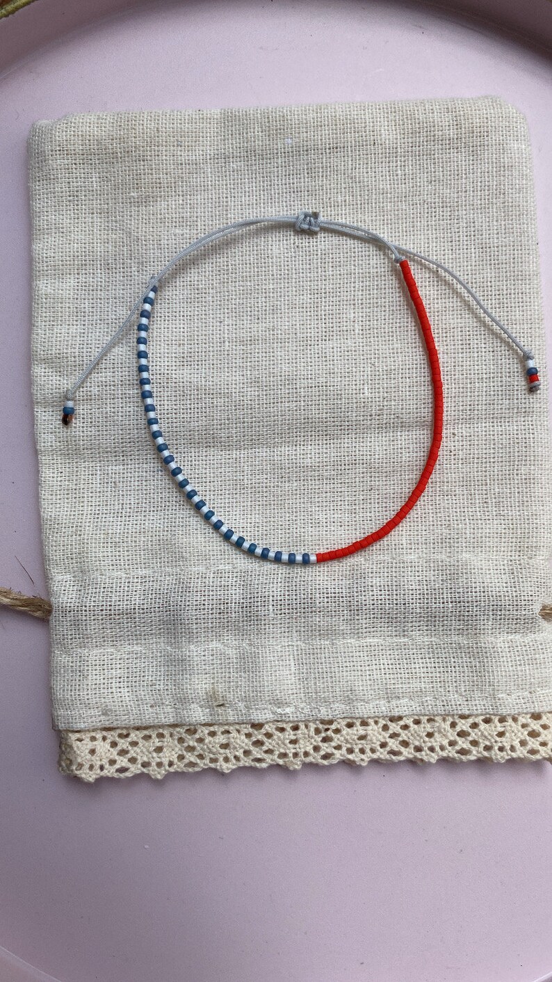 May include: A red and blue beaded bracelet with a white string. The bracelet is on a white fabric background.