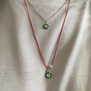 May include: Two necklaces with evil eye pendants. The top necklace is silver with a blue and yellow evil eye pendant. The bottom necklace is red string with a white pearl and a blue and yellow evil eye pendant.