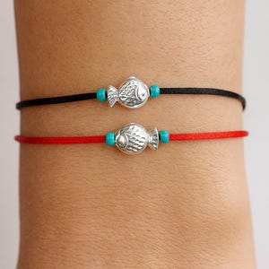 May include: Two bracelets with fish charms. One bracelet has a black cord, the other a red cord. Each bracelet features a silver-colored fish charm and two turquoise beads. The bracelets are worn on a wrist.