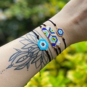 May include: Three beaded bracelets with blue, white, and yellow evil eye charms. The bracelets are on a person's wrist with a black tattoo.