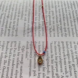 Könnte beinhalten: Ein rotes Armband mit einem goldenen Anhänger, der ein blaues Auge zeigt. Der Anhänger hat die Form einer Träne und ist mit einer goldenen Kette versehen.