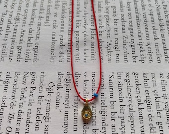 May include: A red string bracelet with a gold charm featuring a blue eye. The charm is in the shape of a teardrop and has a gold chain.