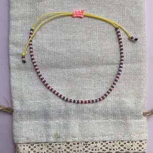 May include: A pink and blue beaded bracelet with a yellow string closure. The bracelet is on a white fabric background.