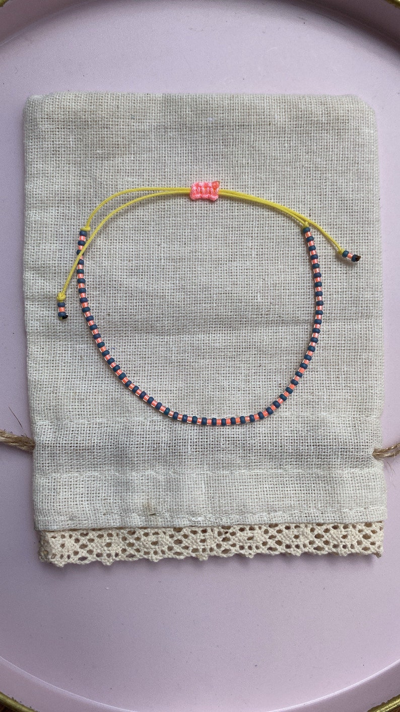 May include: A pink and blue beaded bracelet with a yellow string closure. The bracelet is on a white fabric background.