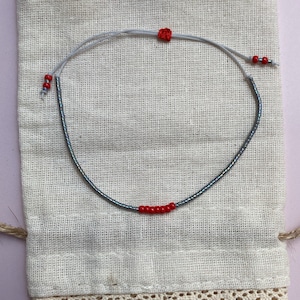 May include: A delicate bracelet made with small gray beads and red accent beads. The bracelet is displayed on a white fabric pouch with lace trim.
