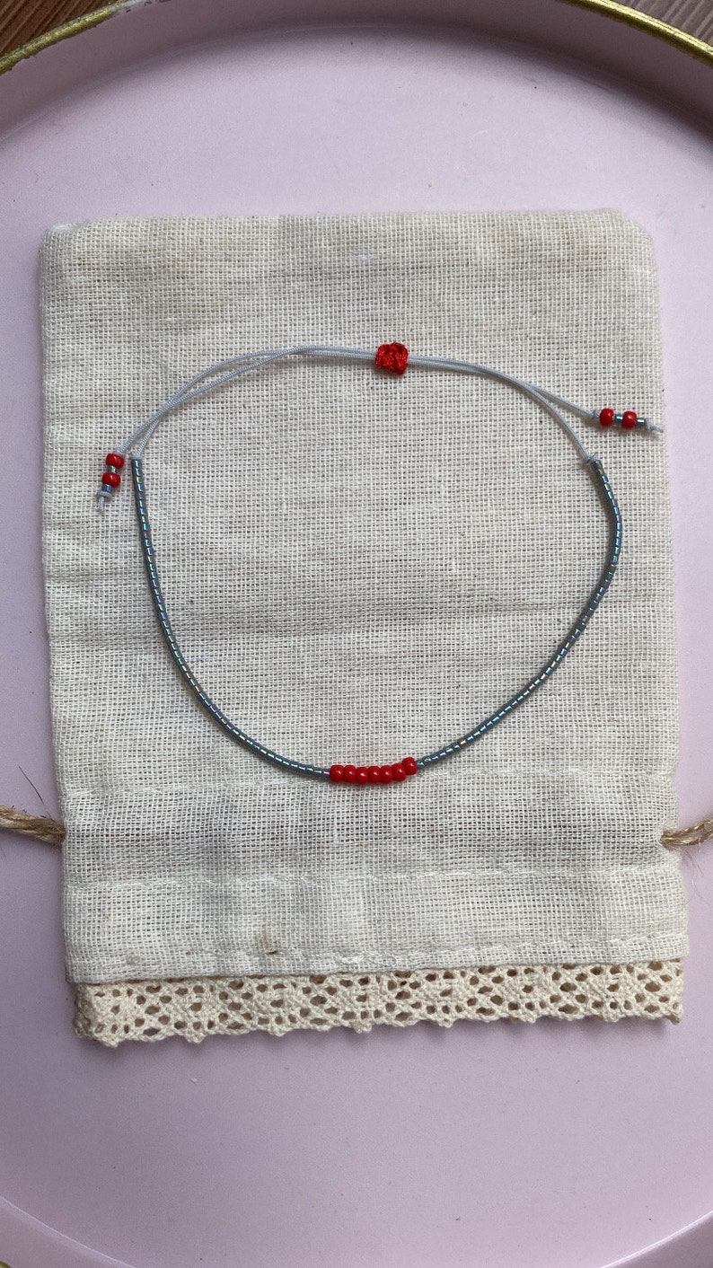 May include: A delicate bracelet made with small gray beads and red accent beads. The bracelet is displayed on a white fabric pouch with lace trim.