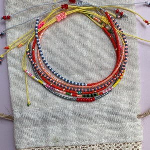 May include: A set of colorful beaded bracelets in a small white fabric bag with lace trim. The bracelets are made with various colors of beads, including red, orange, yellow, blue, and green.