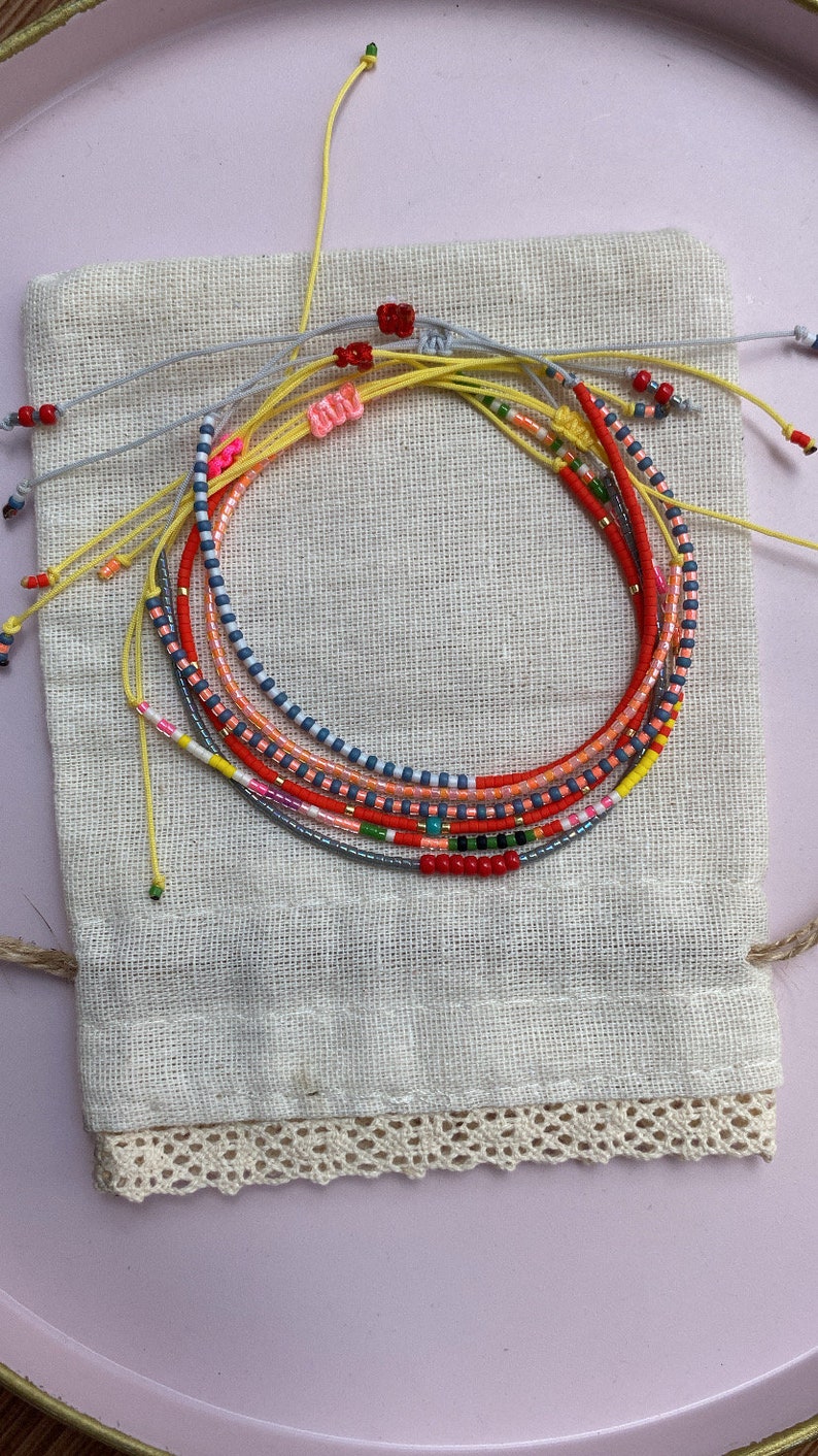 May include: A set of colorful beaded bracelets in a small white fabric bag with lace trim. The bracelets are made with various colors of beads, including red, orange, yellow, blue, and green.