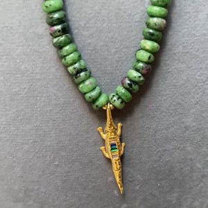 May include: A green gemstone necklace with a gold alligator pendant. The alligator has a row of colorful gemstones on its back.
