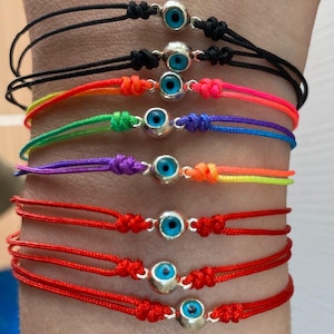 May include: A set of seven colorful string bracelets with silver evil eye charms. The bracelets are layered on a person's wrist, with the colors ranging from black to red.