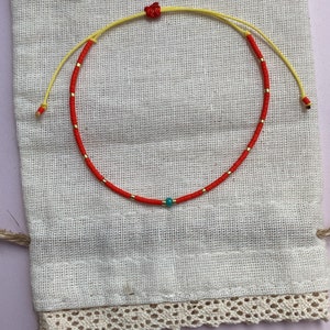 May include: A red and yellow beaded bracelet with a turquoise bead in the center. The bracelet is on a white fabric background.