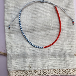 May include: A red and blue beaded bracelet with a white string. The bracelet is on a white fabric background.