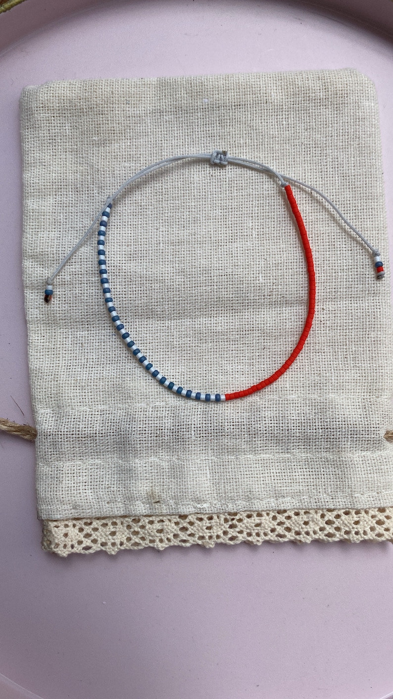 May include: A red and blue beaded bracelet with a white string. The bracelet is on a white fabric background.