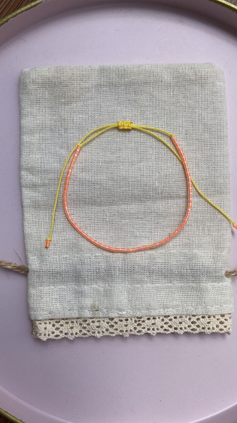 May include: A yellow and orange beaded bracelet on a white fabric pouch with lace trim.