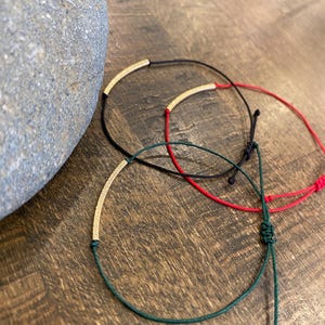 May include: Three minimalist bracelets with gold-coloured curved bars. One bracelet is black, one is red, and one is green. The bracelets are on a wooden surface, with a grey stone in the background. The bracelets are adjustable.