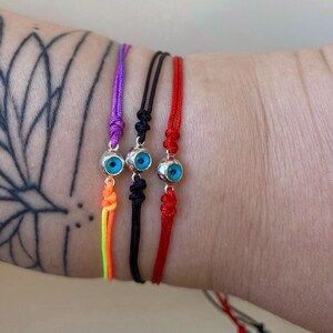 May include: Three adjustable bracelets with an evil eye charm. The bracelets are purple, black, and red. The evil eye charm is silver with a blue eye. The purple bracelet has a rainbow tassel. The bracelets are tied with knots.