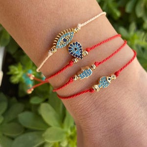May include: Four bracelets with gold accents and blue and white details. One bracelet is off-white, and the other three are red. The bracelets feature various charms, including an eye, a teardrop, a hand, and a fish.