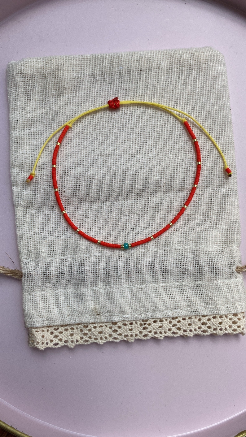 May include: A red and yellow beaded bracelet with a turquoise bead in the center. The bracelet is on a white fabric background.